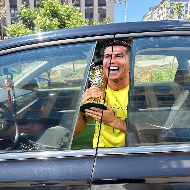 Pode incluir: Uma pessoa segurando um trof&eacute;u de ouro, sorrindo amplamente, vis&iacute;vel atrav&eacute;s da janela de um carro. A pessoa est&aacute; vestindo uma camisa amarela com a palavra "ROYA" impressa. O carro &eacute; preto, e o fundo inclui edif&iacute;cios e vegeta&ccedil;&atilde;o.