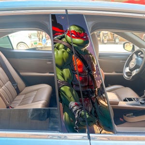 Könnte beinhalten: Ein Autoaufkleber mit einer grünen, muskulösen Zeichentrickschildkröte mit roter Maske, die an Raphael von den Teenage Mutant Ninja Turtles erinnert. Der Aufkleber ist auf dem Fensterrahmen des Autos angebracht, mit einem Stadtlandschaftshintergrund.
