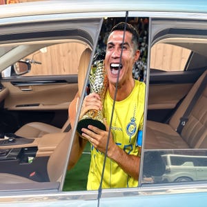 Pode incluir: Uma pessoa com uma camisa amarela segurando um trof&eacute;u dourado, celebrando dentro de um carro. A camisa tem um logotipo e texto. O interior do carro &eacute; vis&iacute;vel, com assentos de couro bege e um exterior azul claro.