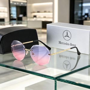 Op de afbeelding: Randloze zonnebril met goudkleurige metalen pootjes en glazen met een roze naar paarse kleurverloop. De bril wordt getoond met een zwarte harde brillenkoker en een zilverkleurige Mercedes-Benz doos met het logo en de tekst op de voorzijde.