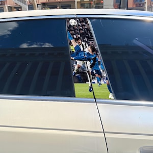 May include: Car window decals featuring a soccer player in a blue uniform performing a bicycle kick, with a soccer ball in the air. The background shows a stadium filled with spectators. The decals are applied to a white car.