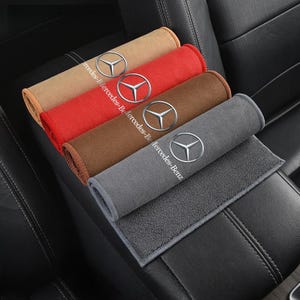 以下が含まれることがあります： ベージュ、赤、茶色、グレーの4色の車のシートベルトカバー。それぞれにシルバーのメルセデス・ベンツのロゴが付いています。カバーは丸められ、黒い革張りの車のシートに展示されています。「Mercedes-Benz」の文字が各カバーに印刷されています。