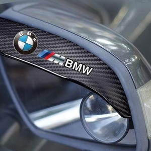 Op de afbeelding: Carbon fiber-textuur autospiegelafdekking met het BMW-logo en de tekst. Het BMW-embleem heeft een blauw-wit cirkelvormig ontwerp, met het BMW-logo en de kleuren van de Duitse vlag. De afdekking is bevestigd aan een autospiegel.