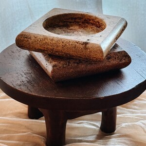 May include: Two stacked square stone trays with a mottled beige and brown finish. Each tray features a smooth, rounded edge and a deep circular indentation in the center. The stone displays natural variations in color and texture throughout.