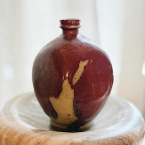 May include: A handcrafted, round, maroon-colored ceramic bottle with a small neck. The bottle has a unique, light-colored, abstract design on its surface. It sits on a light-colored, round, marble-like bowl.