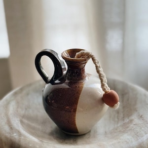 May include: A small, handcrafted ceramic pitcher with a dark brown and white glaze. The pitcher has a looped handle and a rope detail with a small, round wooden bead. The pitcher is displayed on a marble-like, round dish.