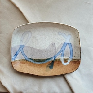 May include: Rectangular ceramic dish with a speckled off-white surface, a brown base, and blue abstract line art. The dish has a rustic, handmade appearance, suitable for serving or display.