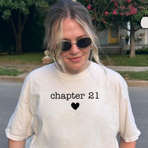 Peut inclure: T-shirt ivoire avec le texte "chapter 21" au-dessus d'un cœur noir. Le t-shirt a des manches courtes et une coupe décontractée. Le modèle porte des lunettes de soleil et un jean bleu clair.