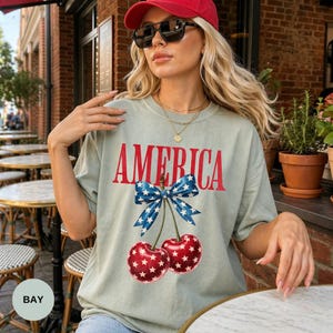 Pode incluir: T-shirt verde claro com a palavra "AMERICA" em vermelho, adornado com um la&ccedil;o azul e cerejas. As cerejas t&ecirc;m padr&otilde;es de estrelas brancas. A pessoa usa um bon&eacute; vermelho e &oacute;culos de sol. A t-shirt &eacute; de estilo casual.