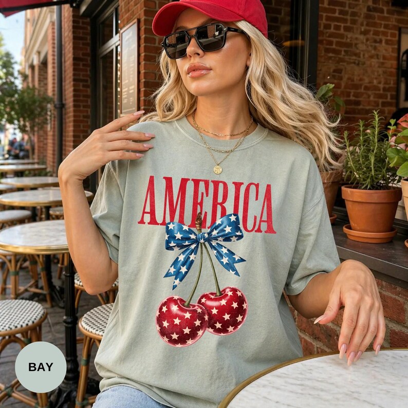 Pode incluir: T-shirt verde claro com a palavra "AMERICA" em vermelho, adornado com um la&ccedil;o azul e cerejas. As cerejas t&ecirc;m padr&otilde;es de estrelas brancas. A pessoa usa um bon&eacute; vermelho e &oacute;culos de sol. A t-shirt &eacute; de estilo casual.