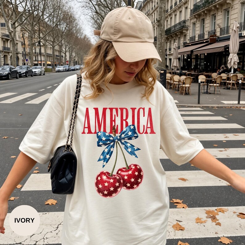 Pode incluir: Camiseta cor marfim com a palavra "AMERICA" em letras vermelhas e um gr&aacute;fico de duas cerejas com um la&ccedil;o azul. As cerejas s&atilde;o vermelhas com padr&otilde;es de estrelas brancas. Um bon&eacute; de beisebol bege &eacute; usado.