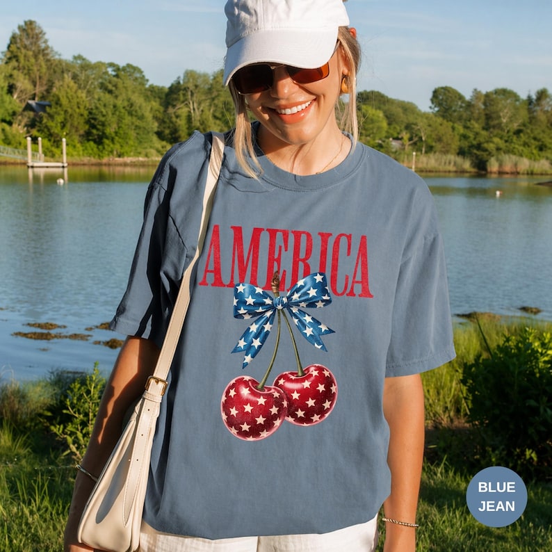 Pode incluir: Camiseta azul jeans com a palavra "AMERICA" em letras vermelhas. Um la&ccedil;o azul com estrelas brancas est&aacute; amarrado a um par de cerejas com padr&otilde;es de estrelas vermelhas e brancas. A camiseta tem um corte folgado.