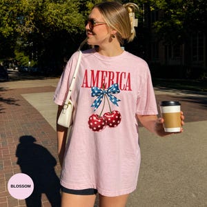 Pode incluir: Uma t-shirt rosa claro com a palavra "AMERICA" em letras vermelhas. O desenho inclui um la&ccedil;o azul com estrelas brancas e duas cerejas com padr&otilde;es de estrelas vermelhas e brancas. A t-shirt &eacute; usada ao ar livre.
