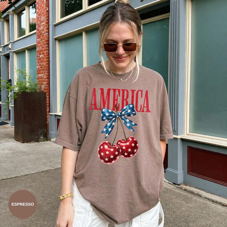 Pode incluir: T-shirt cor espresso com a palavra "AMERICA" em letras vermelhas. Abaixo, um la&ccedil;o azul com estrelas brancas e duas cerejas com padr&otilde;es de estrelas vermelhas e brancas. A camisa &eacute; de estilo casual e oversized.