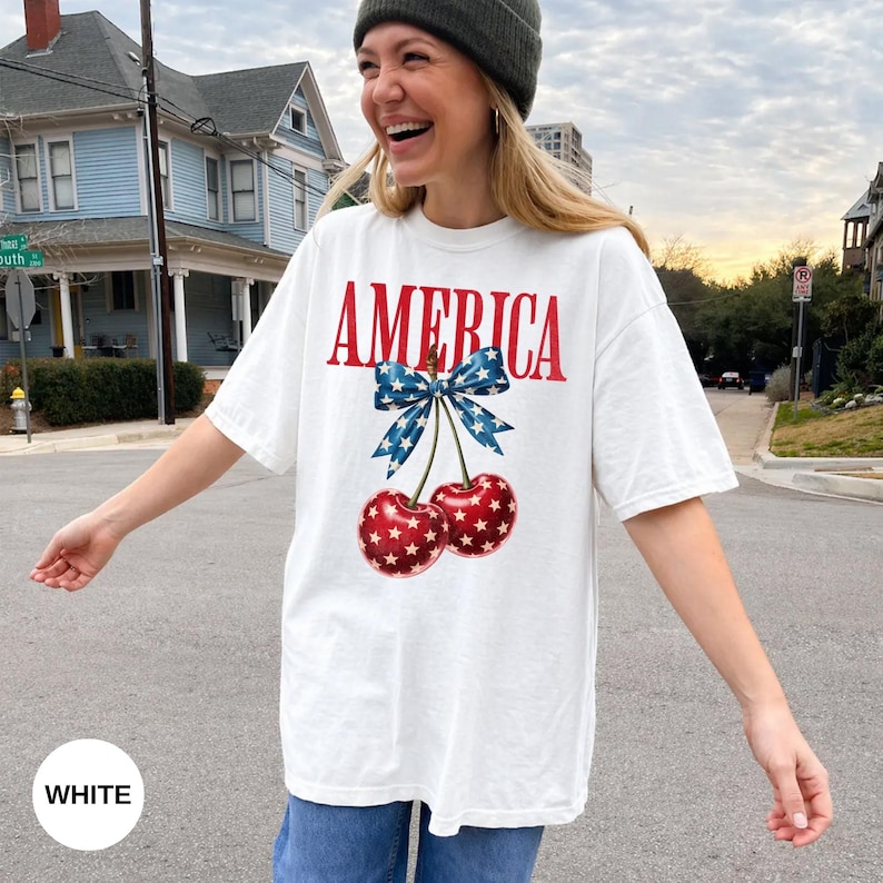 Pode incluir: Camiseta branca com a palavra "AMERICA" em vermelho, um la&ccedil;o azul e duas cerejas. As cerejas s&atilde;o vermelhas com padr&otilde;es de estrelas brancas. A camiseta tem um estilo casual e oversized.
