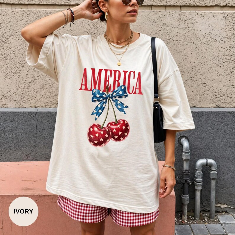 May include: Ivory-colored t-shirt with the word "AMERICA" in red, featuring a cherry design with a blue bow. The cherries are red with white star patterns. The model is wearing red and white checkered shorts.