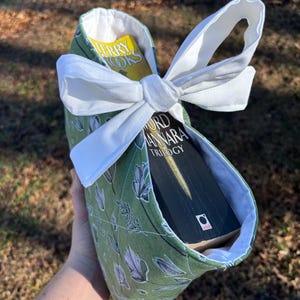 May include: A green fabric book sleeve with a white bow, holding two books. The sleeve has a floral pattern in white and gray. The books are visible inside the sleeve, with titles and author names.