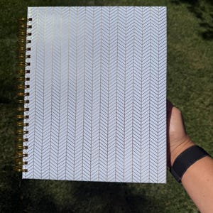 May include: A rectangular notebook featuring a white cover with a repeating gold herringbone pattern. The notebook is bound on the left side with a metallic gold spiral wire coil, allowing the pages to lay flat when opened for writing or sketching.