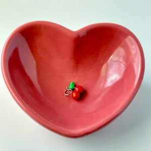 May include: A heart-shaped, red ceramic dish with a pair of cherry earrings. The earrings feature two red cherries and a green leaf. The dish is a solid, glossy red colour.