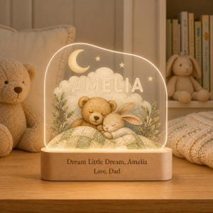 May include: An illuminated acrylic night light featuring a printed illustration of a teddy bear and a rabbit sleeping under a crescent moon and stars. The name AMELIA is printed above the animals. The wooden base reads: Dream Little Dream, Amelia, Love, Dad.