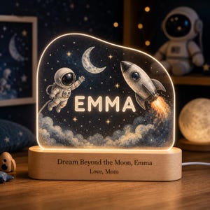 May include: An illuminated acrylic night light featuring a space theme with an astronaut, a crescent moon, and a rocket ship. The name EMMA is displayed in the center. The wooden base is engraved with the text: Dream Beyond the Moon, Emma, Love, Mom.