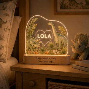 May include: An illuminated acrylic night light featuring a triceratops and a long-necked dinosaur among ferns and eggs. The center displays a heart shape with the name LOLA. The wooden base is engraved with the text Dulces sueños, Lola Con amor, papá.