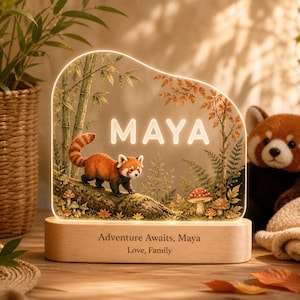 May include: A custom acrylic night light featuring a red panda illustration on a log, bamboo, and mushrooms. The name MAYA is illuminated in white. The light sits on a wooden base engraved with the text: Adventure Awaits, Maya, Love, Family.
