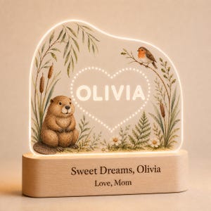 Könnte beinhalten: Ein beleuchtetes Acryl-Nachtlicht mit einem Biber und einem Rotkehlchen in einer Naturszene mit Rohrkolben und Farnen. In der Mitte steht der Name OLIVIA in einem Herz. Der Holzsockel ist mit dem Text Sweet Dreams, Olivia, Love, Mom graviert.