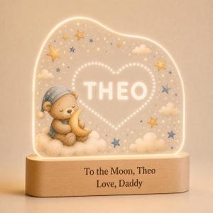 May include: An illuminated acrylic night light featuring a brown teddy bear in a blue nightcap sitting on a cloud and holding a crescent moon. The design includes stars, clouds, and the name THEO inside a heart. The wooden base reads To the Moon, Theo Love, Daddy.
