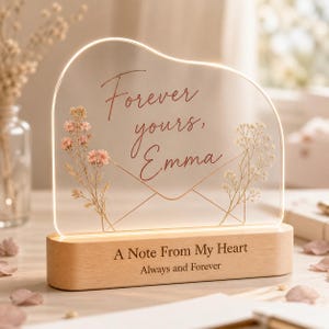 May include: An illuminated acrylic plaque with a wavy top edge, featuring the text "Forever yours, Emma" in script, an envelope outline, and dried pink flowers. The light sits on a wooden base engraved with "A Note From My Heart" and "Always and Forever."