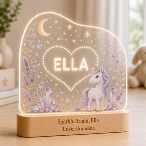 May include: An illuminated acrylic night light featuring a unicorn, crystals, and a crescent moon. The center heart shape displays the name ELLA. The light sits on a wooden base engraved with the text: Sparkle Bright, Ella, Love, Grandma.
