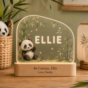 May include: An illuminated acrylic night light featuring a printed illustration of a panda sitting among bamboo stalks. The name ELLIE is displayed in glowing white letters. The light sits on a wooden base engraved with Be Curious, Ellie, Love, Family.