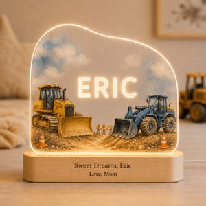 May include: An illuminated acrylic night light featuring a yellow bulldozer and a blue front-end loader on a dirt construction site with orange traffic cones. The name ERIC is displayed in glowing white letters. The wooden base reads Sweet Dreams, Eric, Love, Mom.