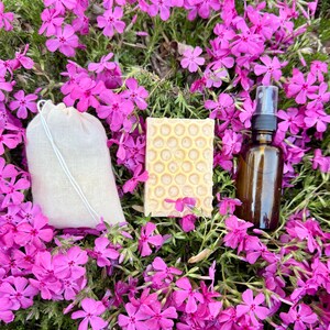May include: A skincare set comprising a beige drawstring bag, a honeycomb-patterned soap bar, and a brown glass spray bottle. The items are set amongst vibrant pink flowers, creating a fresh, organic aesthetic.