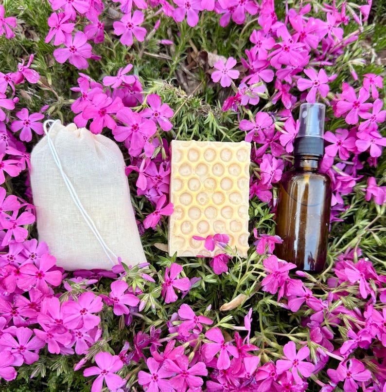 May include: A skincare set comprising a beige drawstring bag, a honeycomb-patterned soap bar, and a brown glass spray bottle. The items are set amongst vibrant pink flowers, creating a fresh, organic aesthetic.