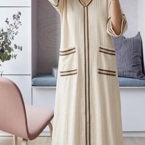 Könnte beinhalten: Ein cremefarbenes, langärmliges Kleid mit V-Ausschnitt und zwei Vordertaschen. Das Kleid hat dunkelbraune Verzierungen am Ausschnitt, vorne und um die Taschen und Ärmel. Das Kleid ist aus leichtem Stoff gefertigt.