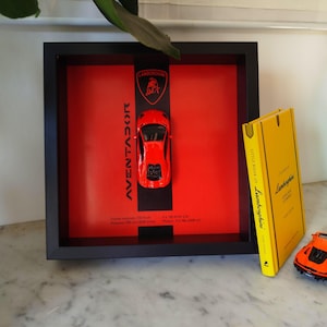 May include: A framed red Lamborghini Aventador toy car with the Lamborghini logo and text. A yellow book titled "Little Book of Lamborghini" and an orange toy car are also present.