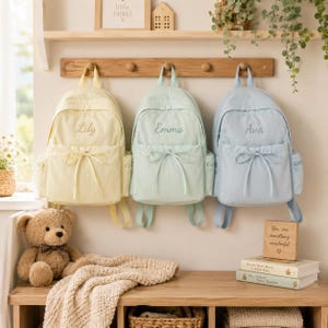 May include: Three backpacks in pastel yellow, mint green, and light blue, each featuring a front pocket with a decorative bow, side pockets, and embroidered names Lily, Emma, and Ava on the front. The backpacks hang from a wooden wall rack.