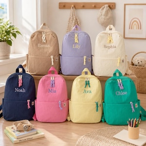 May include: A collection of seven backpacks in various solid colors including tan, lavender, cream, navy blue, pink, yellow, and green. Each backpack features a front pocket, a top handle, and a name embroidered on the front in a contrasting color.