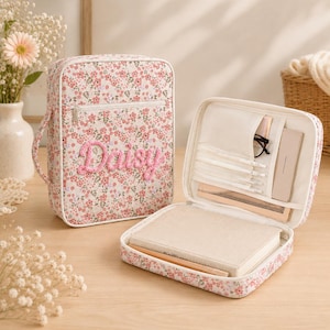 Puede incluir: Un estuche organizador con estampado floral y el nombre Daisy bordado en rosa en la parte frontal. Cuenta con cierre de cremallera, un asa lateral y un interior con múltiples bolsillos, ranuras para bolígrafos y un cuaderno beige en su interior.