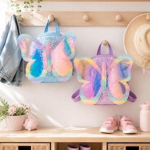 Puede incluir: Dos mochilas con forma de mariposa, una azul y otra morada. Ambas bolsas presentan alas holográficas iridiscentes con un borde de degradado arcoíris y un cuerpo con lentejuelas. Cada mochila tiene un asa superior y correas ajustables.