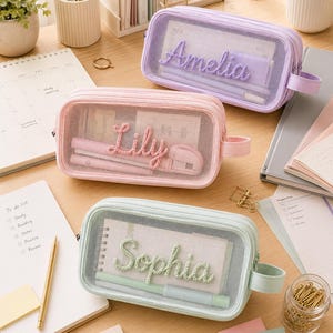 May include: Three rectangular pencil cases in pastel lavender, pink, and mint green. Each case features a transparent mesh front panel, a side handle, and a name embroidered in matching yarn: Amelia, Lily, and Sophia. The cases contain stationery.