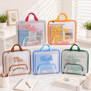 May include: Five rectangular, transparent storage bags with colored fabric trim and handles. Each bag features a personalized name in raised script lettering and a smaller front pocket with the text "good lucky" containing pens and pencils.