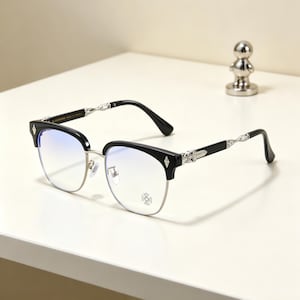 Puede incluir: Gafas negras y plateadas con diseño de media montura. Las monturas presentan una sección superior negra con detalles en forma de diamante y bordes inferiores plateados. Las patillas tienen detalles decorativos plateados y terminales negros. Un pequeño emblema de cruz plateada está en el borde inferior.