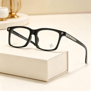 Könnte beinhalten: Schwarze rechteckige Brille mit silbernen Kreuz- und Blumenakzenten. Die Brille hat einen dicken Rahmen und klare Gläser. Die Bügel weisen dekorative silberne Details auf. Die Brille wird auf einer weißen Box präsentiert.
