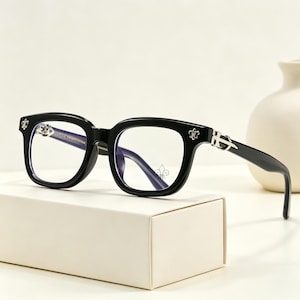 May include: Black square-framed eyeglasses with clear lenses. The frames have silver fleur-de-lis and decorative accents on the temples. The glasses are displayed on a white box, with a white vase in the background.