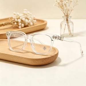 May include: Clear eyeglasses with a square frame design. The temples and hinges feature silver-colored decorative accents. The lenses are clear, and the frame has a small, circular emblem on the lower right lens. The glasses are displayed on a wooden tray.
