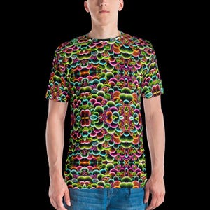 May include: A short-sleeve t-shirt featuring a vibrant, repeating fractal pattern of rounded shapes in neon shades of pink, green, orange, and blue. The design covers the entire garment, which is paired with blue denim jeans against a black background.