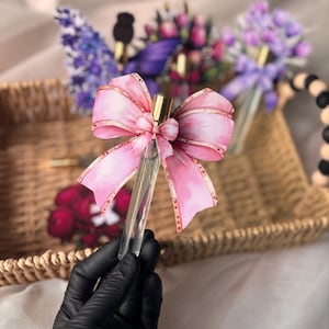 Op de afbeelding: Een heldere glazen parfumfles met een roze strik. De strik heeft een aquarelontwerp met gouden accenten. De fles wordt vastgehouden door een gehandschoende hand. Andere bloemstukken staan op de achtergrond, in een gevlochten mand.