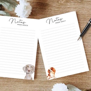 May include: Two white notepads with lined pages and dog illustrations. One notepad has a Weimaraner, the other a Cavalier King Charles Spaniel. The text "Notes from Bruce" and "Notes from Keely" are at the top. A black pen rests on the right.
